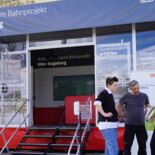 Drei Menschen stehen vor dem Infomobil des Bahnprojekts Ulm-Augsburg und unterhalten sich