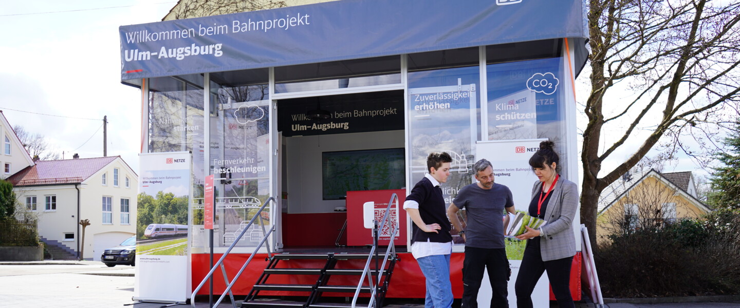 Drei Menschen stehen vor dem Infomobil des Bahnprojekts Ulm-Augsburg und unterhalten sich